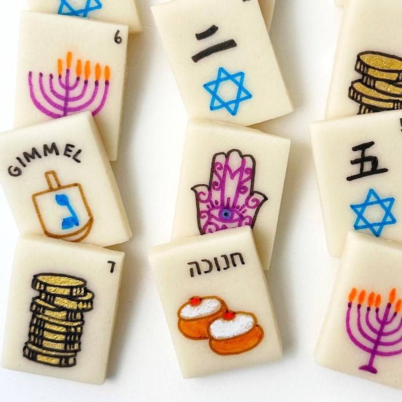 Op de afbeelding: Ivoor-kleurige tegels met diverse handgetekende illustraties en cijfers. Ontwerpen omvatten een menora, een dreidel, munten, een Hamsa-hand en Hebreeuwse en Japanse karakters. Een unieke set voor spellen of display.