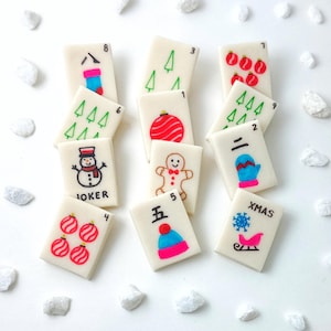 May include: A collection of rectangular tiles with Christmas-themed illustrations. The tiles feature images of ornaments, a snowman, a gingerbread man, and a snowflake with a sleigh. Some tiles have numbers and the word "JOKER" and "XMAS".