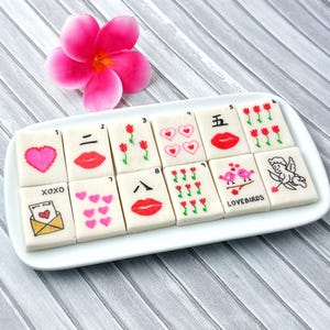 May include: A white rectangular platter holds decorated cookies with Valentine's Day themes. Designs include hearts, lips, tulips, and an envelope. A pink flower sits above the platter. The cookies are arranged on a gray and white wooden surface.
