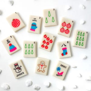 May include: A collection of Christmas-themed Mahjong tiles. Each tile features a number and a festive illustration, including ornaments, a mitten, Christmas trees, a stocking, a snowman, a gingerbread man, and a sleigh with the word "XMAS".