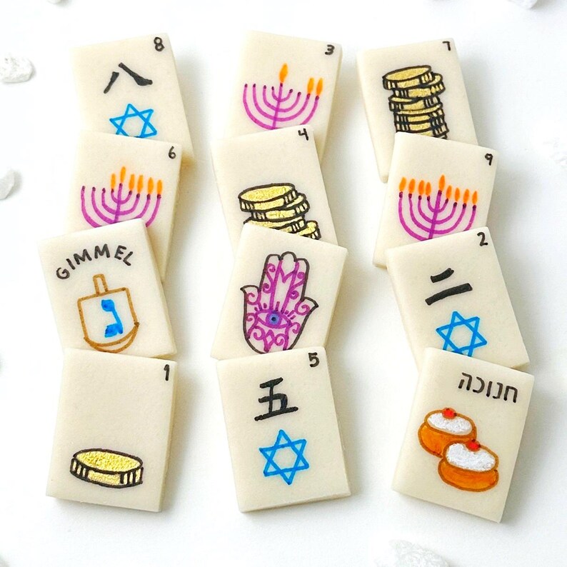 Op de afbeelding: Een verzameling rechthoekige, cr&egrave;mekleurige speltegels met diverse handgetekende afbeeldingen en cijfers. De illustraties omvatten een menora, munten, een Davidster, een dreidel, een Hamsa-hand en tekst in meerdere talen. De tegels liggen op een witte ondergrond.