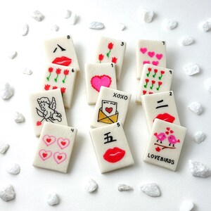 May include: A collection of white rectangular tiles with various Valentine's Day-themed illustrations. Designs include hearts, lips, flowers, an envelope, and a cupid. The tiles are scattered on a white surface with small white decorative stones.