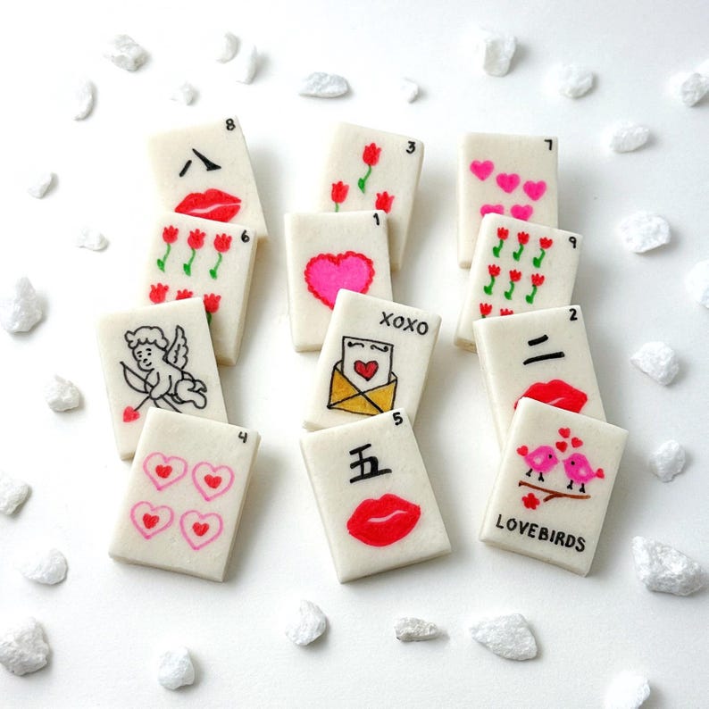 May include: A collection of white rectangular tiles with various Valentine's Day-themed illustrations. Designs include hearts, lips, flowers, an envelope, and a cupid. The tiles are scattered on a white surface with small white decorative stones.