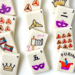 Op de afbeelding: Een verzameling rechthoekige, gebroken witte koekjes versierd met kleurrijke Purim-thema ontwerpen. De koekjes tonen afbeeldingen van kronen, maskers en driehoeken, samen met cijfers en het woord "Purim".