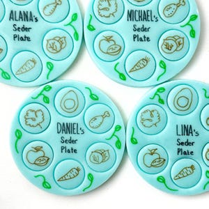 May include: Four blue and white Seder plates with the names Alana's, Michael's, Daniel's, and Lina's written on them. Each plate has six sections with different symbols representing the Seder plate items.
