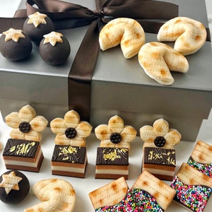 Peut inclure: Assortiment de friandises décoratives, comprenant des pâtisseries en forme de fleurs, des biscuits de fortune et des carrés enrobés de chocolat avec des accents de feuilles d'or. Les friandises sont disposées sur une surface blanche avec une boîte cadeau argentée et un ruban marron en arrière-plan.