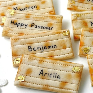 May include: Close-up of several rectangular, light-brown cookies with horizontal lines and gold leaf accents. Some cookies have names written in black ink, while one says "Happy Passover". A festive treat for a special occasion.
