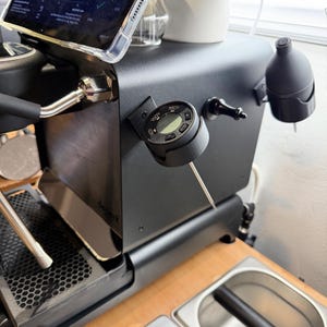 Puede incluir: Máquina de espresso negra con termómetro digital adjunto. Una tableta que muestra un gráfico está montada encima de la máquina. La máquina tiene un portafiltro de acero inoxidable y una bandeja de goteo. La configuración está sobre una superficie de madera.