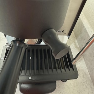 Peut inclure: Gros plan sur une machine à expresso noire avec une buse vapeur en acier inoxydable. La machine est dotée d'un bac d'égouttement noir et d'un porte-filtre noir. La marque "Cent" est visible.