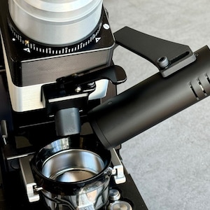 Könnte beinhalten: Eine schwarze Kaffeemühle mit einem silbernen Trichter und einem schwarzen Siebträger. Die Mühle hat ein Zifferblatt mit Zahlen und einen Hebel. Der Siebträger ist an einem schwarzen Auslauf befestigt.