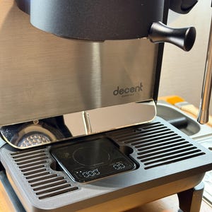 Op de afbeelding: Een espresso machine van roestvrij staal met een zwarte bovenkant en een digitale weegschaal. De machine heeft de tekst "decent espresso" op de voorkant. De weegschaal geeft cijfers weer. De machine staat op een houten oppervlak.