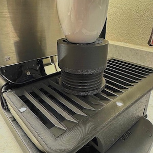 May include: A black metal drip tray with a grid pattern and a white ceramic cup sitting on top of a black metal stand. The stand has a black rubber base. The text "Sage" is visible on the tray.