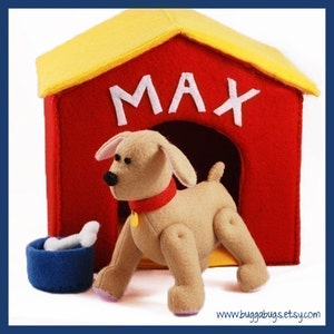 Puede incluir: Una casa para perros de fieltro rojo con un techo amarillo y el nombre "MAX" escrito en la parte delantera. Un perro de fieltro marrón con un collar rojo está de pie frente a la casa para perros.