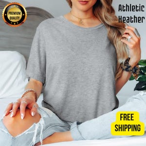 K&ouml;nnte beinhalten: Ein meliertes graues T-Shirt mit Rundhalsausschnitt. Das Shirt besteht aus einem weichen Material. Die Worte "Athletic Heather" sind in Schwarz gedruckt. Ein goldenes Siegel mit den Worten "Premium Quality" befindet sich in der oberen linken Ecke.
