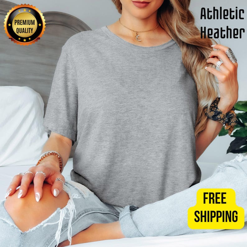 K&ouml;nnte beinhalten: Ein meliertes graues T-Shirt mit Rundhalsausschnitt. Das Shirt besteht aus einem weichen Material. Die Worte "Athletic Heather" sind in Schwarz gedruckt. Ein goldenes Siegel mit den Worten "Premium Quality" befindet sich in der oberen linken Ecke.