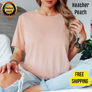 K&ouml;nnte beinhalten: Ein meliertes pfirsichfarbenes T-Shirt mit kurzen &Auml;rmeln. Das Shirt ist einfarbig und hat einen Rundhalsausschnitt. Die Worte "Heather Peach" sind in Schwarz gedruckt. Ein goldfarbenes Premium-Qualit&auml;tsabzeichen befindet sich in der oberen linken Ecke. Die Worte "FREE SHIPPING" stehen in einer gelben Box.
