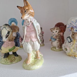 May include: A ceramic fox figurine standing on a green base, wearing a light blue coat, a pink waistcoat, and white breeches. The fox has orange fur and is positioned in the foreground, with other animal figurines visible in the background.