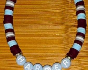 May include: A beaded bracelet featuring flat, circular clay discs in alternating colors of maroon, light blue, white, and gray. The bracelet includes six round, light blue beads with the white lettered text HARPER centered at the bottom.