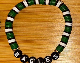 May include: A beaded bracelet with green, white, and black disc-shaped beads. The word "EAGLES" is spelled out in black letter beads. The bracelet is on a wooden surface.