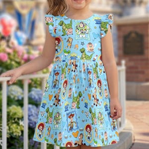 Puede incluir: Una ni&ntilde;a peque&ntilde;a con un vestido azul claro con un estampado de personajes de Toy Story. El vestido tiene mangas con volantes y un cuello redondo. La ni&ntilde;a sonr&iacute;e frente a una valla blanca y un fondo borroso.
