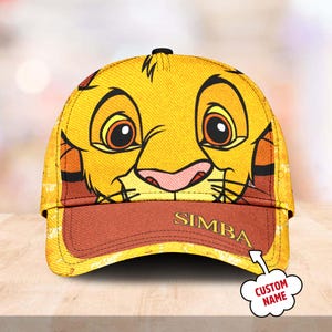 May include: Yellow baseball cap featuring a cartoon image of Simba from The Lion King. The cap has a red brim with the text "SIMBA" printed on it. The text "CUSTOM NAME" is written in a speech bubble.