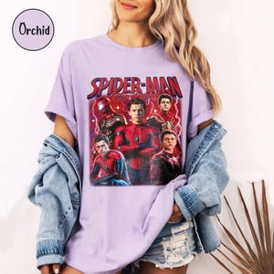 K&ouml;nnte beinhalten: Ein helllila T-Shirt mit einer Spider-Man-Grafik und dem Text "Spider-Man: Peter Parker". Das Design zeigt mehrere Bilder von Spider-Man in verschiedenen Posen und Kost&uuml;men. Das T-Shirt hat kurze &Auml;rmel.