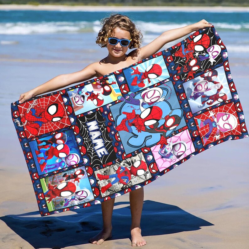 Puede incluir: Una toalla de playa rectangular con un collage de ilustraciones de Spider-Man y Spider-Gwen en rojo, azul y blanco. La toalla tiene un borde azul oscuro e incluye la palabra "NAME" en blanco. El fondo es una playa de arena.