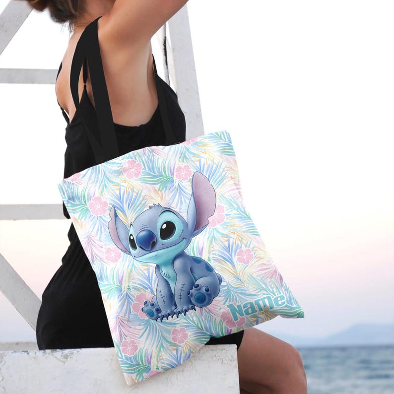 May include: A blue tote bag with a cartoon character, Stitch, with large ears and big eyes. The bag has a colourful floral and leaf pattern in pastel colours. The word "Name" is printed on the bag.