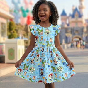 Puede incluir: Un vestido azul claro con un estampado de personajes de dibujos animados. El vestido tiene un cuello redondo y mangas cortas con volantes. La falda se ensancha desde la cintura. El fondo est&aacute; borroso, pero parece ser un parque tem&aacute;tico.