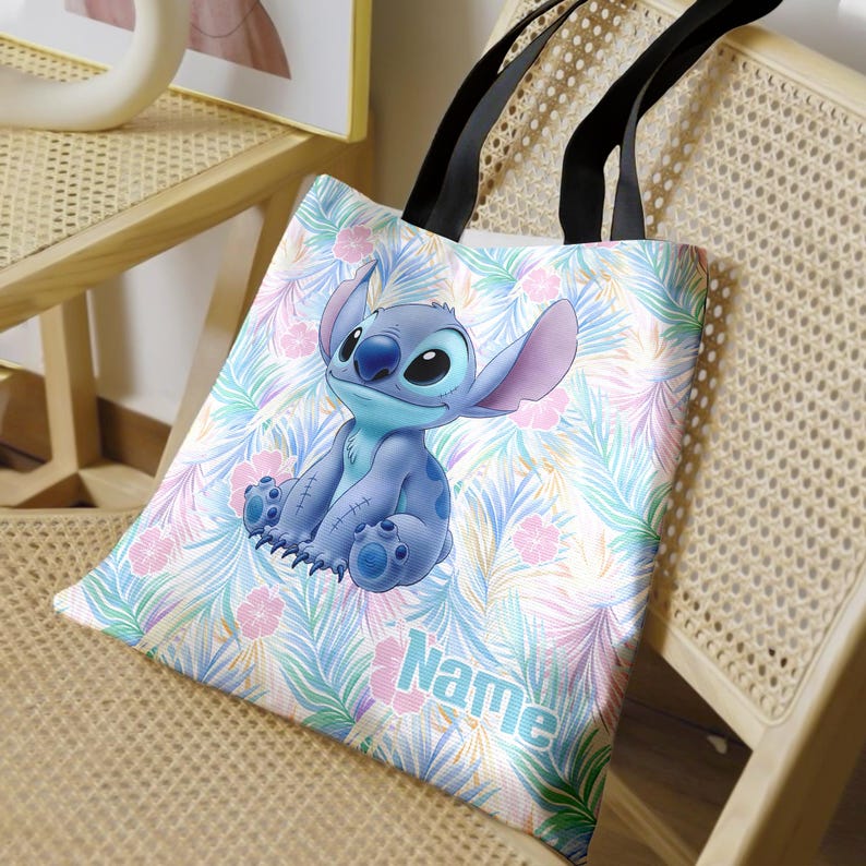 May include: A white tote bag with a blue cartoon character on a tropical leaf and flower pattern. The bag has black handles and the word "Name" printed on the front. The bag is suitable for carrying items.