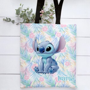 May include: A blue tote bag with a cartoon character, Stitch, on a tropical floral pattern. The bag has black handles and the word "Name" in light blue. The background features pink flowers and green leaves.