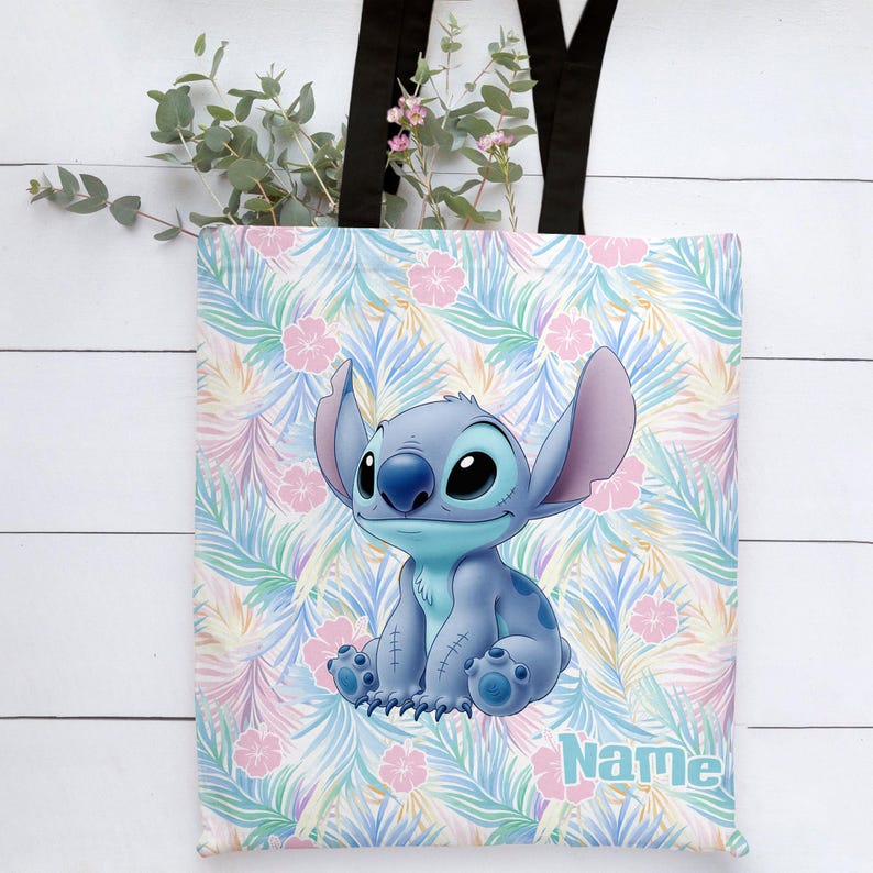 May include: A blue tote bag with a cartoon character, Stitch, on a tropical floral pattern. The bag has black handles and the word "Name" in light blue. The background features pink flowers and green leaves.