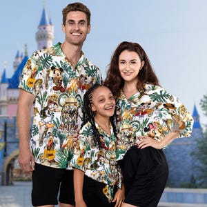 May include: Three people wearing matching white button-down shirts with a colourful print featuring Disney characters and jungle animals. The shirts have short sleeves and are paired with black shorts. The text "Animal Kingdom" is visible on the shirts.