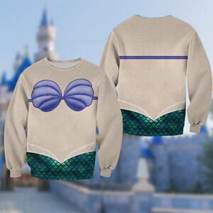 Op de afbeelding: Een beige sweatshirt met een zeemeerminontwerp. De voorkant toont een blauwe schelpenbeha en een groene schaalpatroon onderkant. De achterkant heeft een blauwe streep en een groen schaalpatroon.