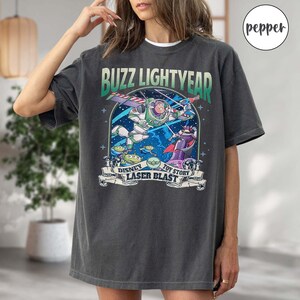 Puede incluir: Una camiseta gris oscuro con un gr&aacute;fico de estilo vintage de Buzz Lightyear de Toy Story. El gr&aacute;fico presenta a Buzz en su traje espacial, volando en una nave espacial con el texto "Buzz Lightyear", "Disney", "Toy Story" y "Laser Blast".