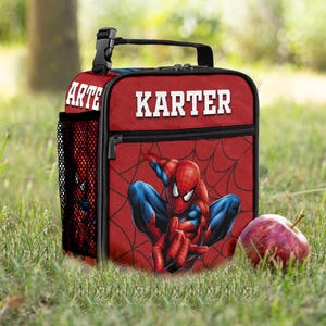 May include: A red rectangular lunch bag featuring a graphic of a superhero in a blue and red suit with a black spiderweb pattern. The name KARTER is printed in white block letters on the front. It includes a black handle and a side mesh pocket.