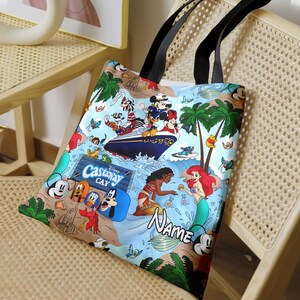 May include: A blue and white tote bag with a Disney Castaway Cay theme. The bag features Mickey Mouse, Minnie Mouse, and other Disney characters. The bag also has the text "Disney Castaway Cay" and "Name".
