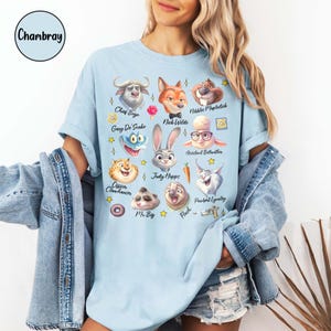 May include: Light blue t-shirt featuring cartoon illustrations of characters from Zootopia, including Chief Bogo, Nick Wilde, and Judy Hopps. The shirt has short sleeves and a crew neck. The word "Chambray" is in a circle in the upper left corner.