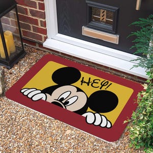 May include: A rectangular doormat featuring Mickey Mouse peeking over a yellow background with the word "HEY!" in black. The mat has a red border and is placed on a gravel surface near a door.