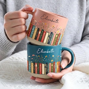 May include: Two ceramic mugs with speckled finishes, one in dusty pink and one in teal. Both feature the name Elizabeth in script, with a row of colourful illustrated books at the base and delicate wildflowers extending upwards from the book spines.