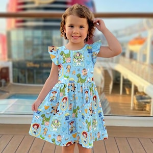 Puede incluir: Un vestido azul claro con un estampado de Toy Story. El vestido tiene cuello redondo, mangas con volantes y un corpi&ntilde;o ajustado que fluye hacia una falda acampanada. El estampado incluye personajes como Buzz Lightyear, Woody y Rex. El vestido lo lleva una ni&ntilde;a.