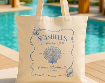 Op de afbeelding: Crèmekleurige draagtas met blauwe tekst en schelpmotieven. De tekst luidt "Seashells & Wedding Bells" met "Olivia's Bachelorette, New York" eronder. Een grote schelpgrafiek staat in het midden van de tas, met het jaar "2025" rechts afgedrukt.
