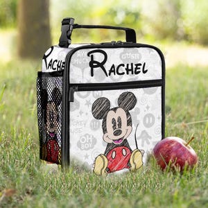 May include: White lunch bag with a Mickey Mouse sketch design and the name "Rachel" in black. It has a black handle, zip, and side pocket. The background features Mickey Mouse icons. An apple sits on the grass.