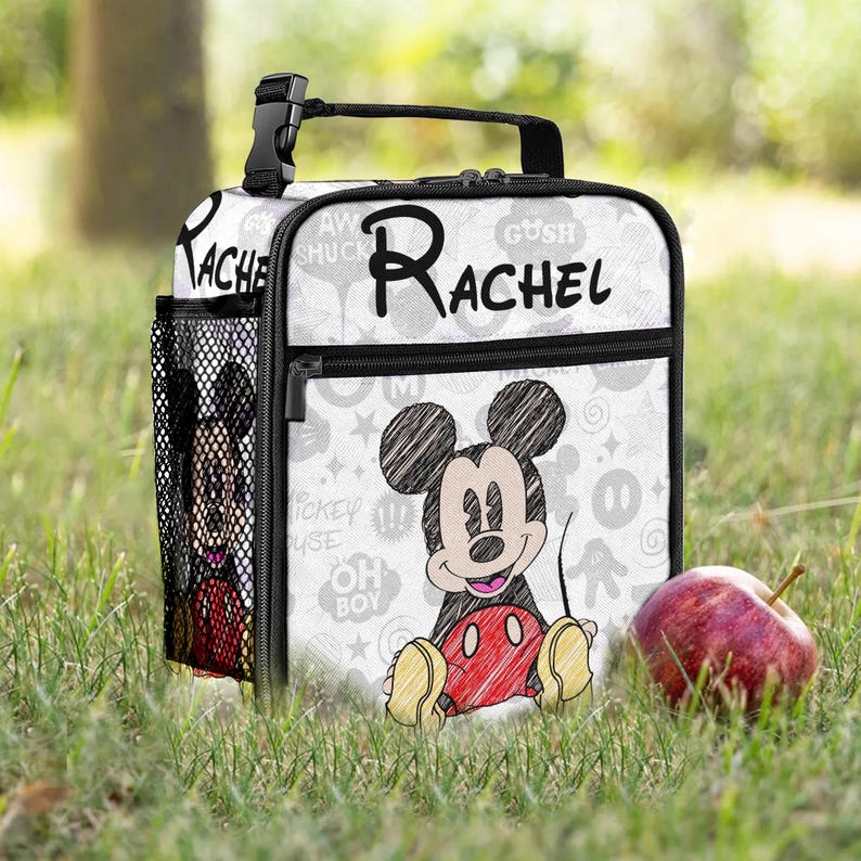 May include: White lunch bag with a Mickey Mouse sketch design and the name "Rachel" in black. It has a black handle, zip, and side pocket. The background features Mickey Mouse icons. An apple sits on the grass.