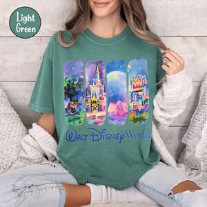 Puede incluir: Camiseta verde claro con cuatro dise&ntilde;os rectangulares verticales de acuarela con el tema de Walt Disney World. Los dise&ntilde;os incluyen un &aacute;rbol, un castillo con fuegos artificiales, una escena a la luz de la luna y un edificio colorido. Las palabras "Walt Disney World" est&aacute;n impresas en azul.