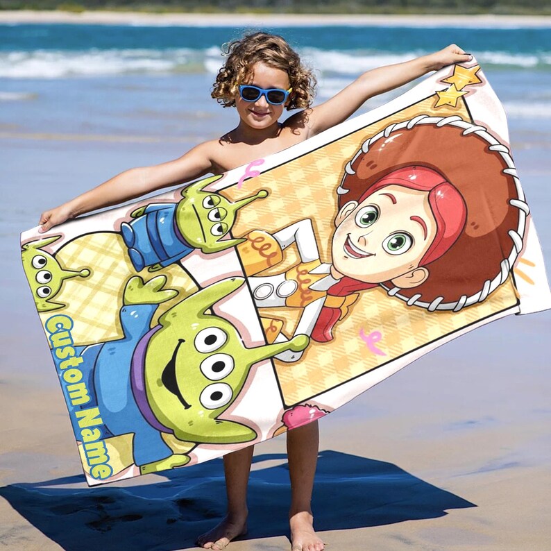 Puede incluir: Una toalla de playa rectangular con personajes de dibujos animados de Toy Story. La toalla tiene un fondo claro con un patr&oacute;n repetitivo. Los personajes incluyen a los Little Green Men y a Jessie la vaquera. Las palabras "Custom Name" est&aacute;n impresas en la toalla.