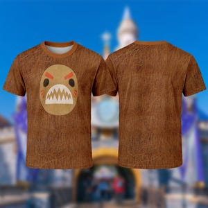 A brown t-shirt with a cartoon monster design on it, set against a blurred background of a castle or palace.
