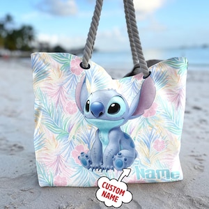 May include: A beach tote bag with a cartoon character design, set against a tropical floral pattern in pastel shades. The bag has grey rope handles. The word "Name" is printed on the bag.
