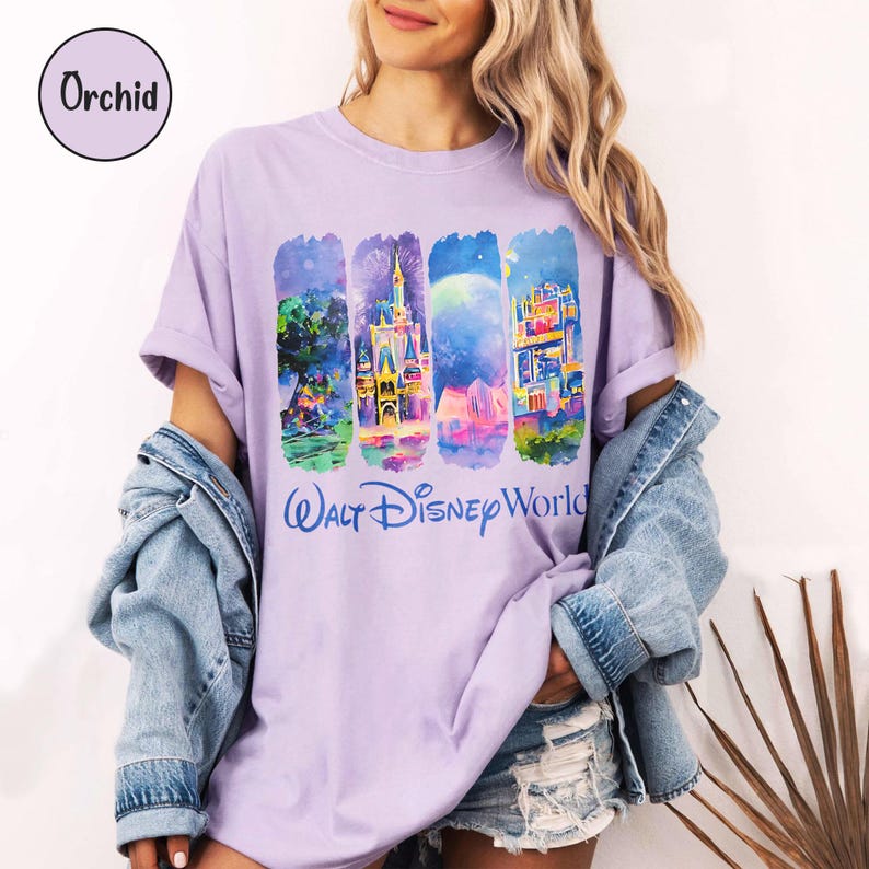 Puede incluir: Una camiseta lila con un gr&aacute;fico colorido de escenas de Walt Disney World. El gr&aacute;fico incluye im&aacute;genes de un castillo, fuegos artificiales y otras atracciones del parque. El texto "Walt Disney World" est&aacute; impreso en azul. La camiseta tiene mangas enrolladas.