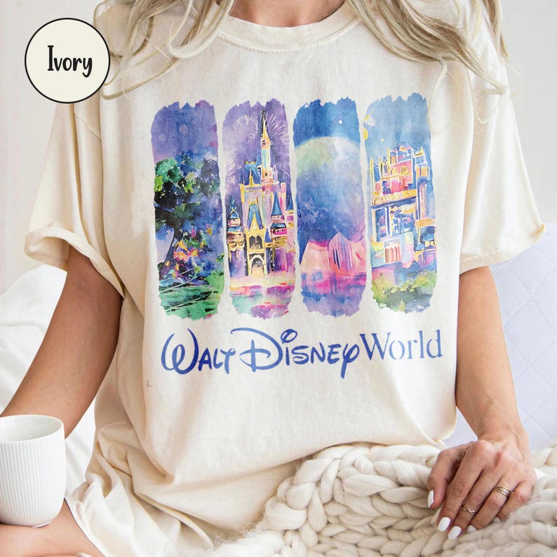 Puede incluir: Camiseta de color crema con un dise&ntilde;o de acuarela de los monumentos de Walt Disney World. El dise&ntilde;o incluye im&aacute;genes de un castillo, fuegos artificiales y otras atracciones. Las palabras "Walt Disney World" est&aacute;n impresas en azul.
